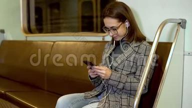一个年轻的女人用耳机听音乐，她手里拿着一部手机在地铁火车上。 这个女孩正在写信